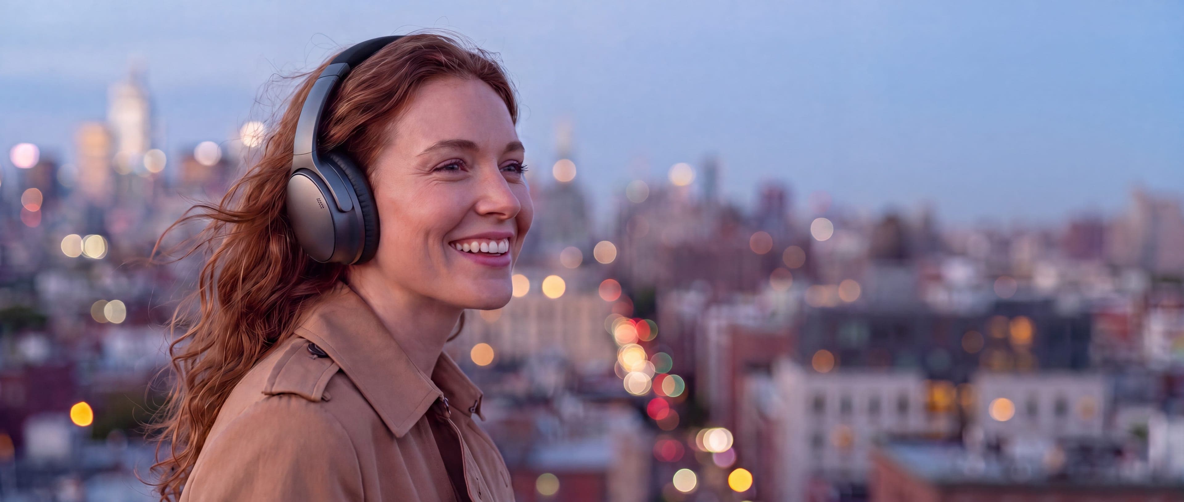 Smiling woman on a city rooftop at dusk wearing premium black noise-cancelling headphones